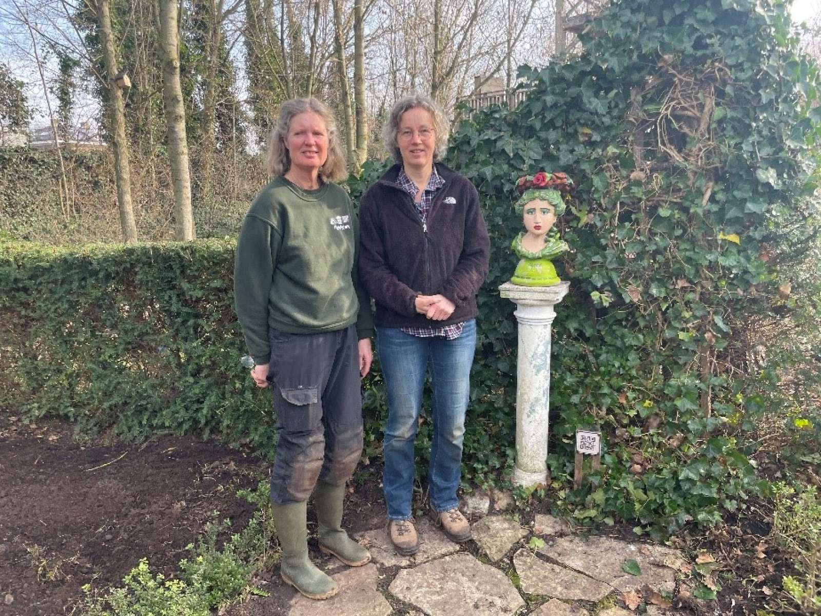 Karin Kok (links) en Judith Uiterwijk naast een standbeeldje op een pilaar, met haag en bomen er achter