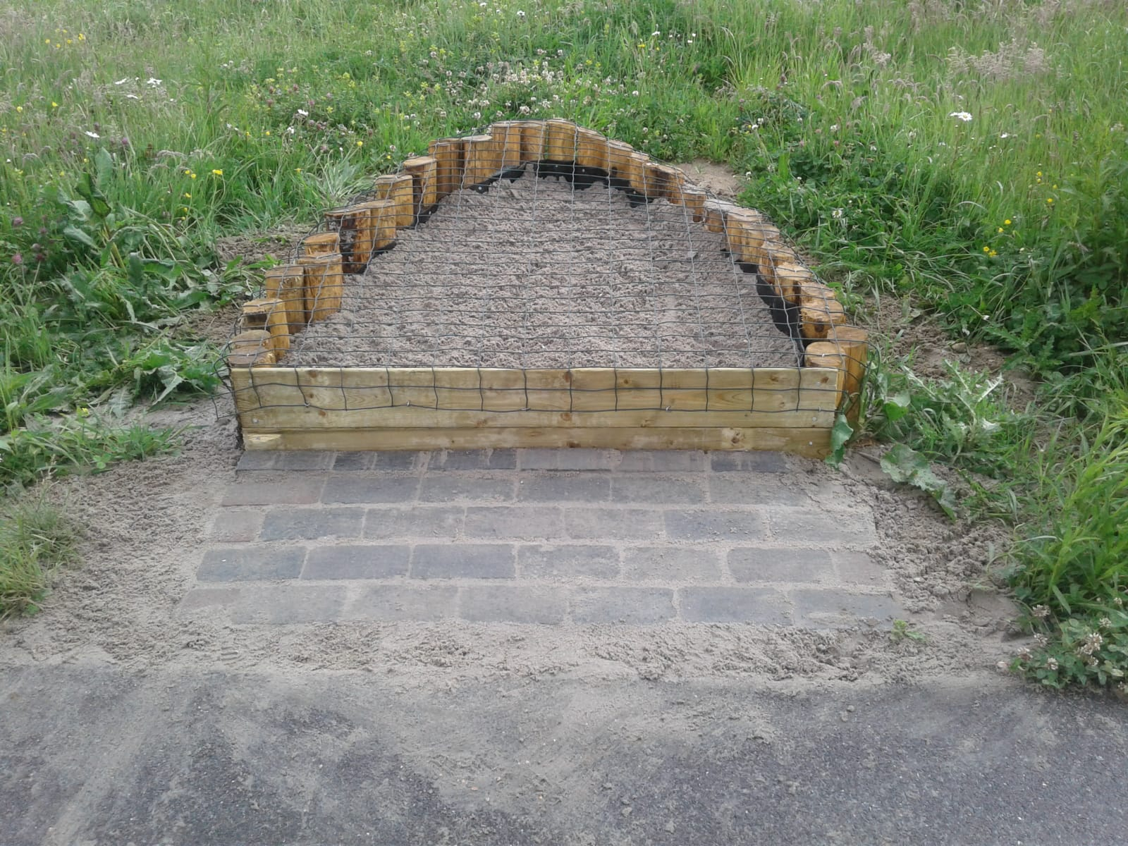 Een aangelegde bijenheuvel: hoop zand omgeven door paaltjes en een plank, met gaas tegen gravende katten e.d.
