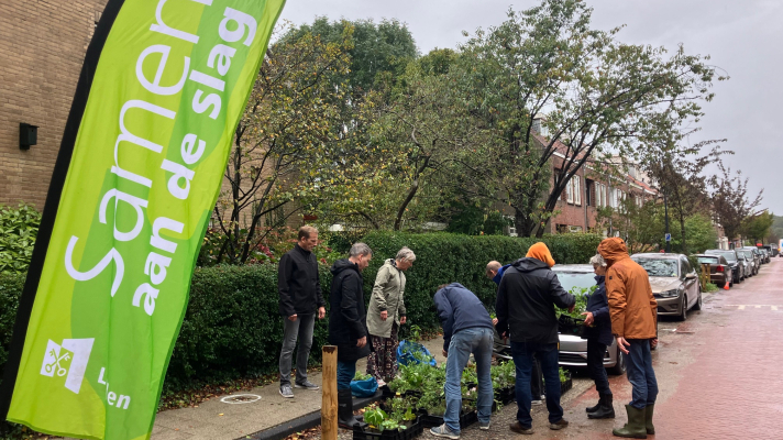 buurtbewoners zoeken plantjes uit voor een Samen aan de Slag - plantactie. Vooraan een beachflag met 'Samen aan de Slag'